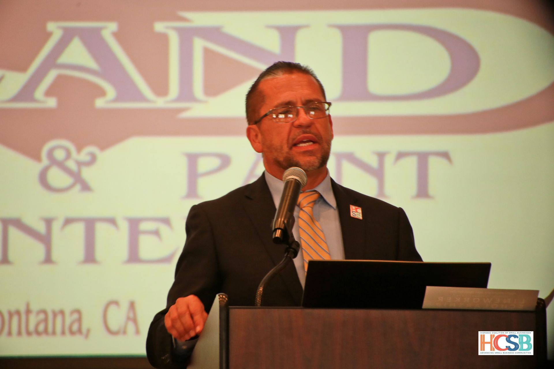 A man in a suit and tie stands at a podium speaking into a microphone