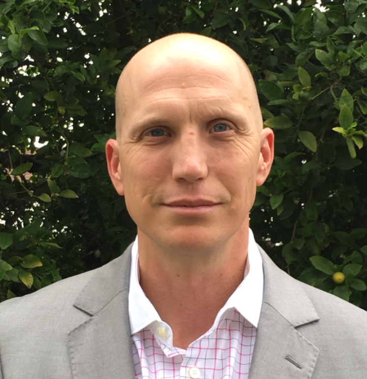 A bald man in a suit and plaid shirt stands in front of a tree