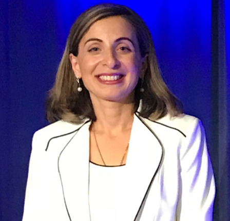 A woman in a white jacket is smiling in front of a blue curtain