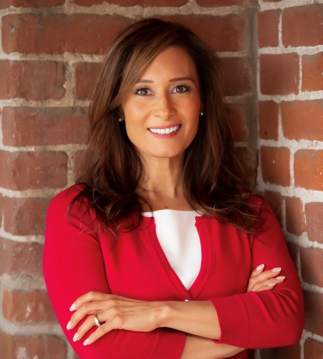 A woman in a red sweater is standing in front of a brick wall with her arms crossed