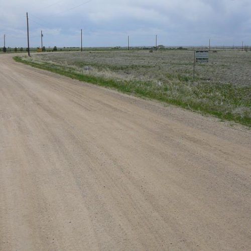 Dirt road leads through a grassy field, under a cloudy sky. Power lines visible.