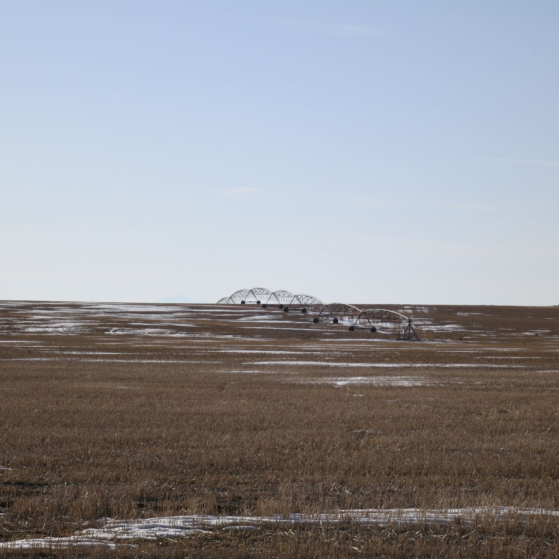 Brown field with patches of snow under a light blue sky; a distant, low-lying structure is in the center.