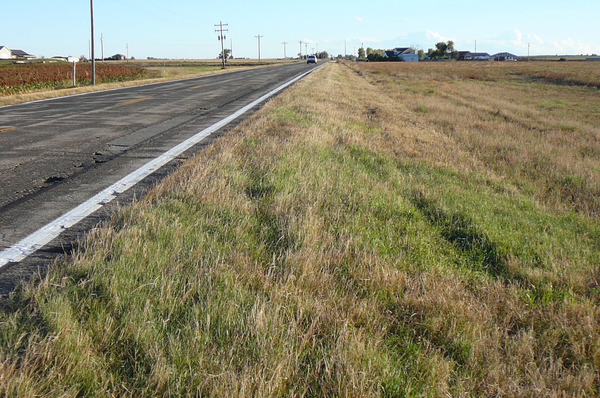Road with a cracked asphalt surface and a grassy verge, under a sunny sky.