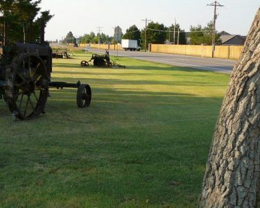 Rusty antique farm equipment on a grassy lawn next to a road, wooden fence, and trees.