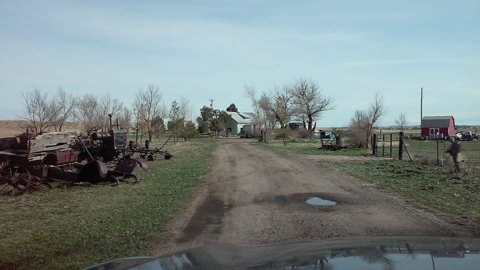 Dirt road leading to a rural farmhouse, flanked by fields and old farm equipment, under a cloudy sky.
