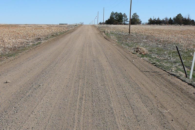 Dirt road stretching into the distance between fields under a blue sky, power lines on the right.