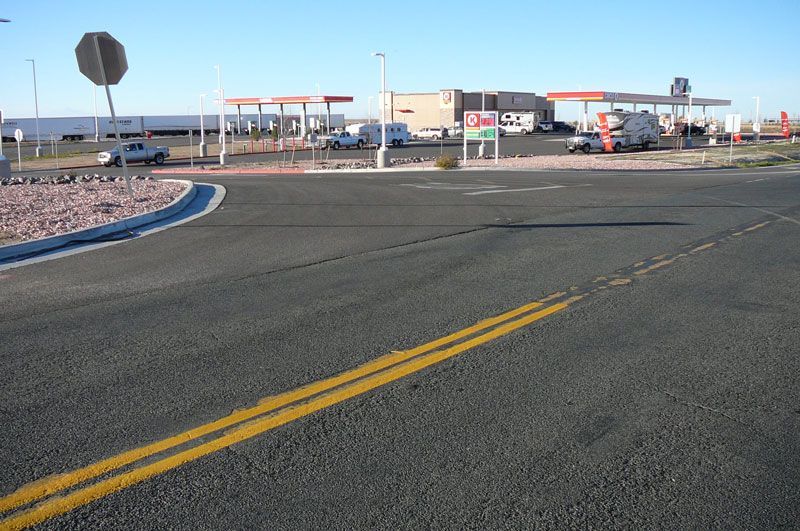 Paved road with double yellow lines, leading to a gas station with several vehicles and a stop sign.