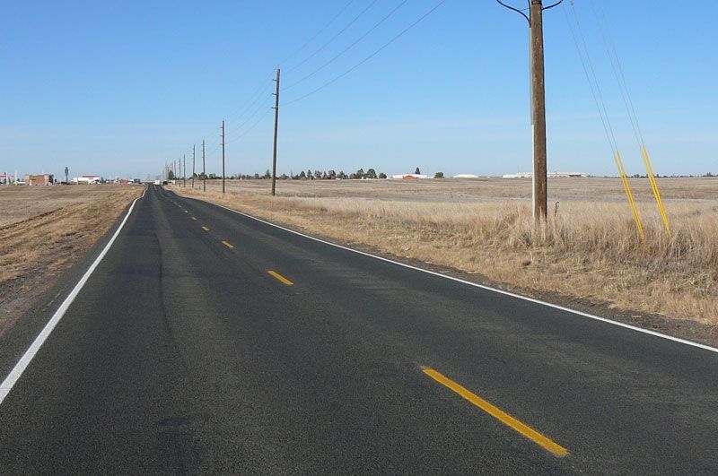 Paved road through a flat, brown field under a clear blue sky. Power poles line the roadside.