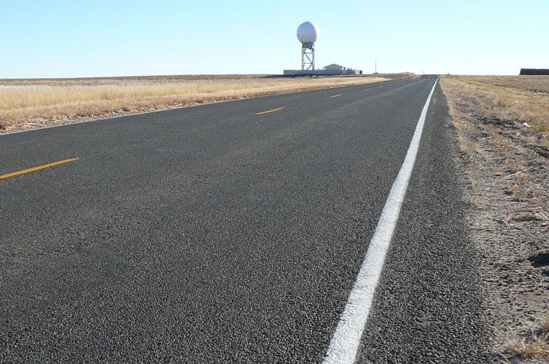 Road stretching towards a water tower on a flat, brown landscape.