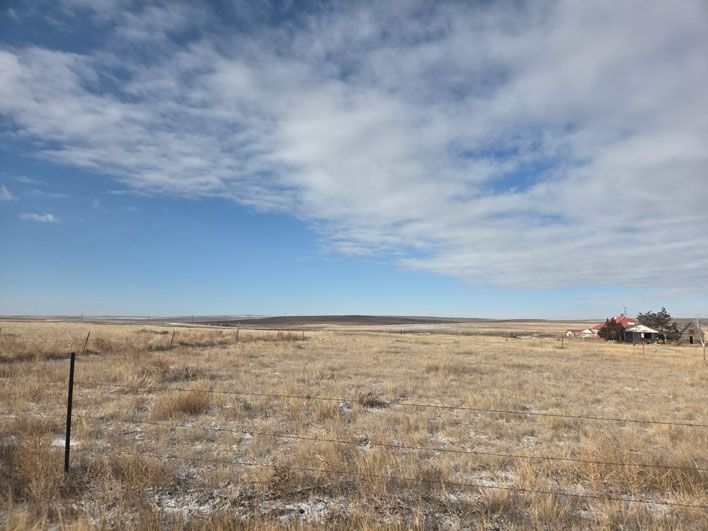 Vast, brown grassland under a blue sky with scattered clouds. A dilapidated building is visible on the right.