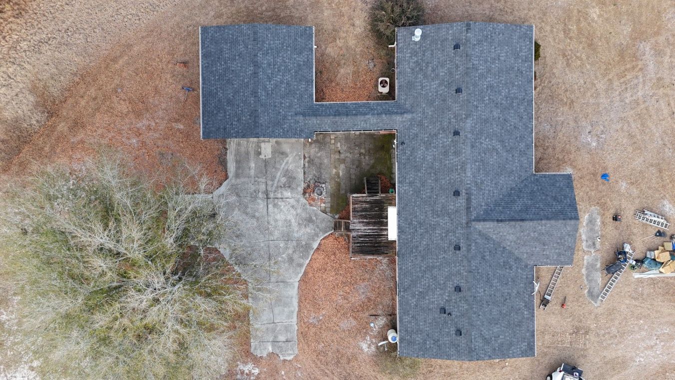 Aerial view of a house with a dark roof, driveway, and surrounding yard with trees and dirt.