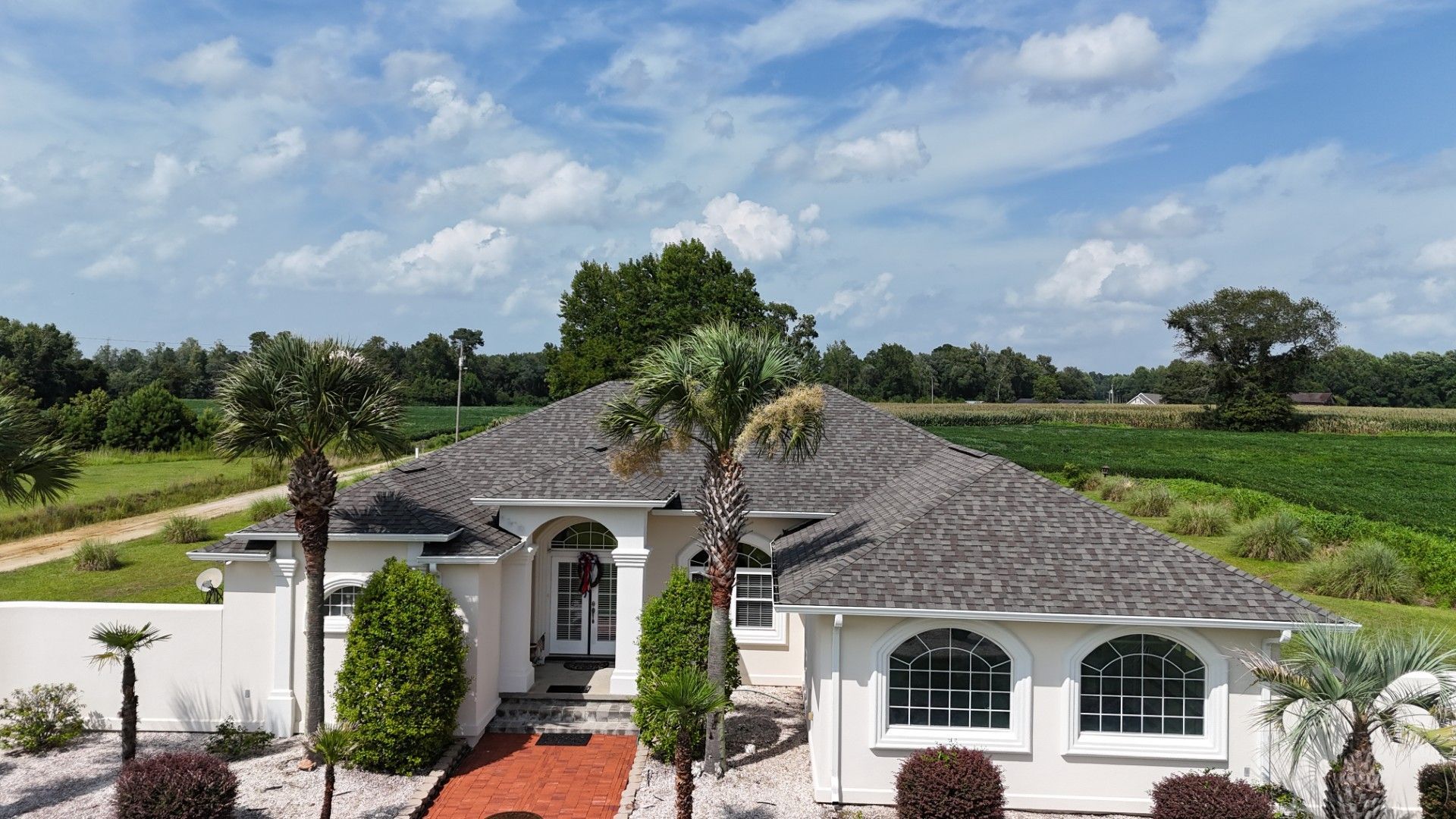 White house with gray roof amid green fields under a partly cloudy sky, viewed from above