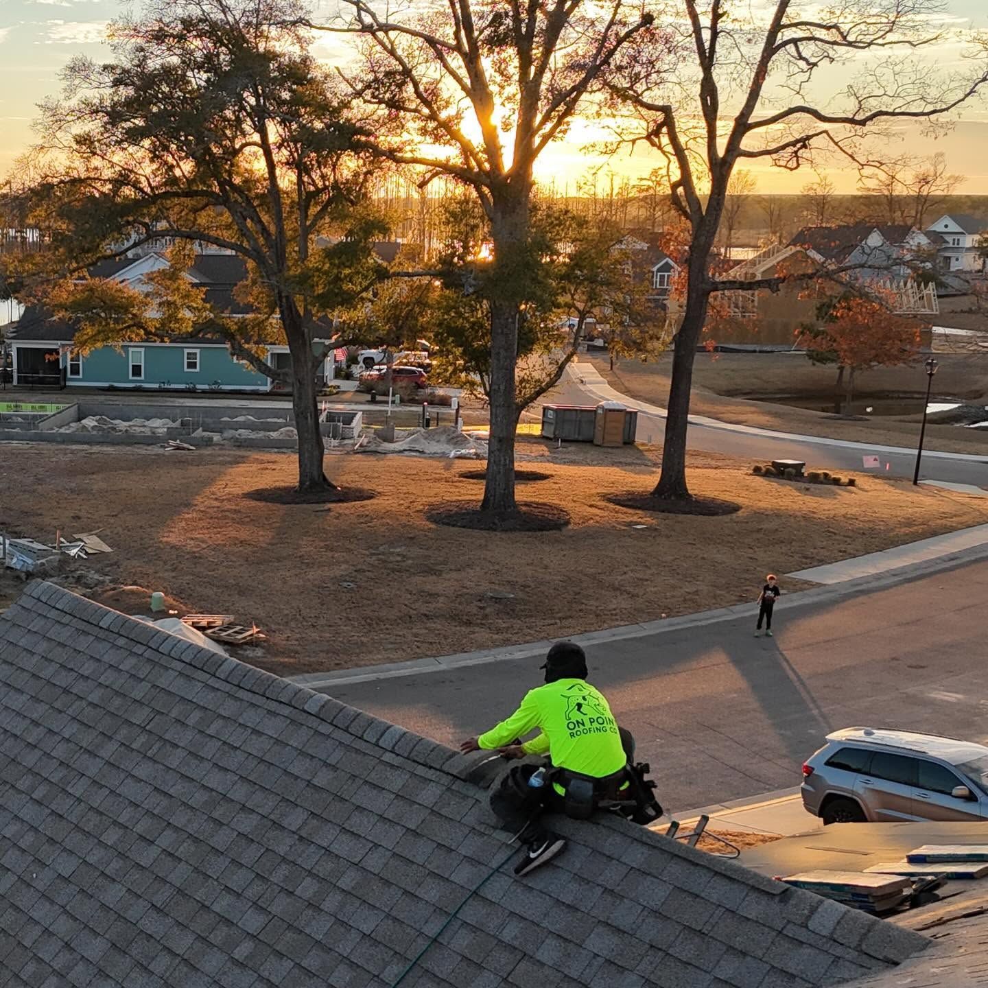 Worker in a neon jacket on a roof at sunset, overlooking a snowy suburban street with bare trees.