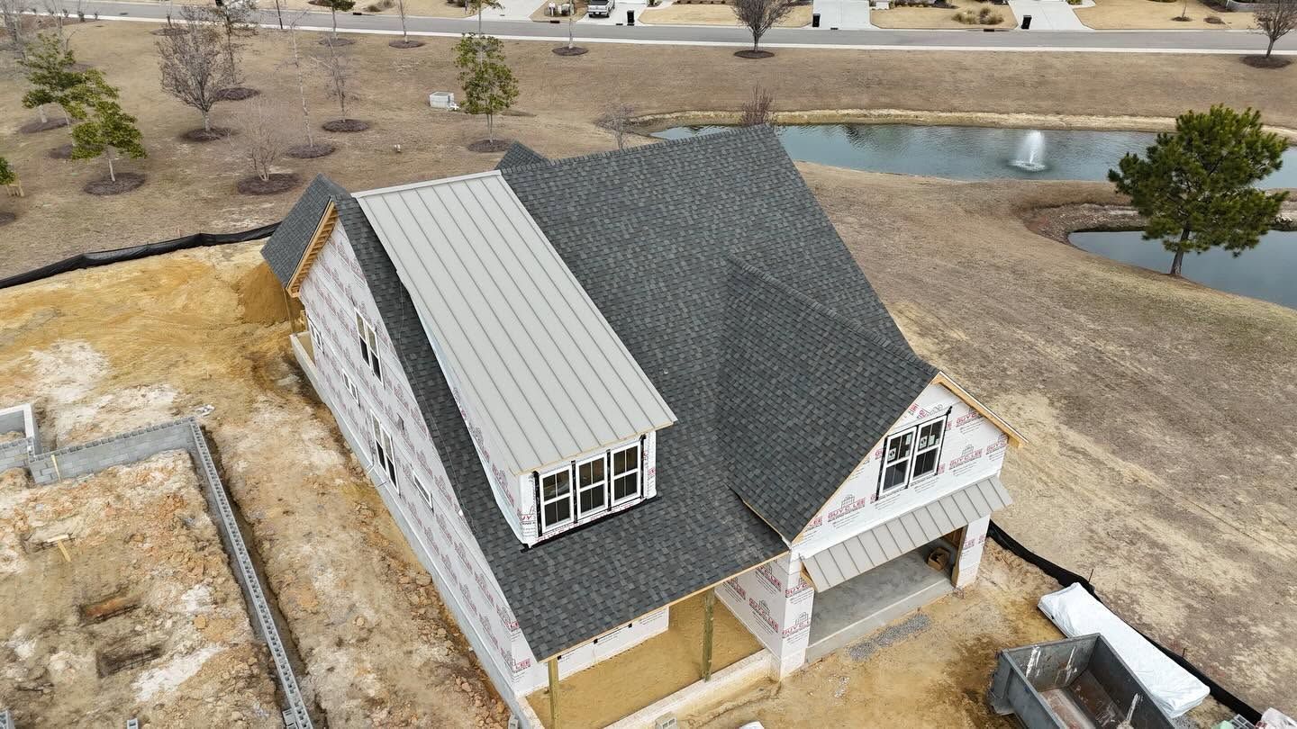 Aerial view of a gray-roofed house with a light metal-roof addition near a pond in a dry suburban area