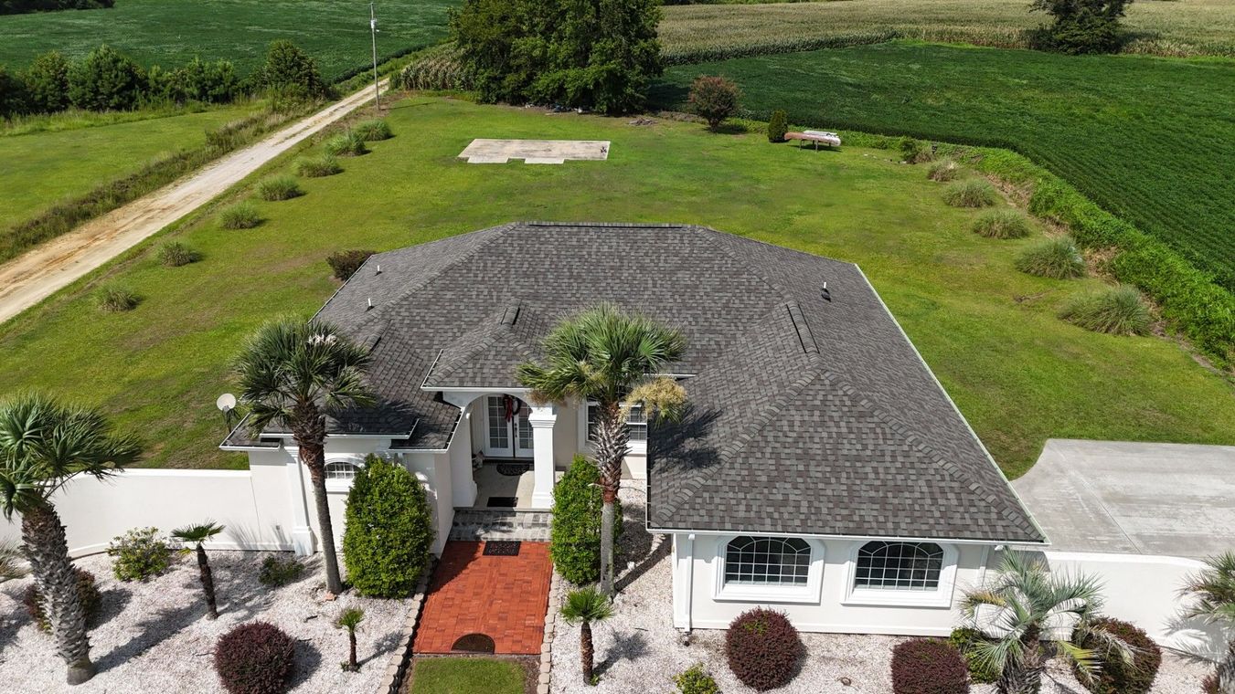 Aerial view of a white house with a dark roof, red front walkway, and green fields surrounding it.
