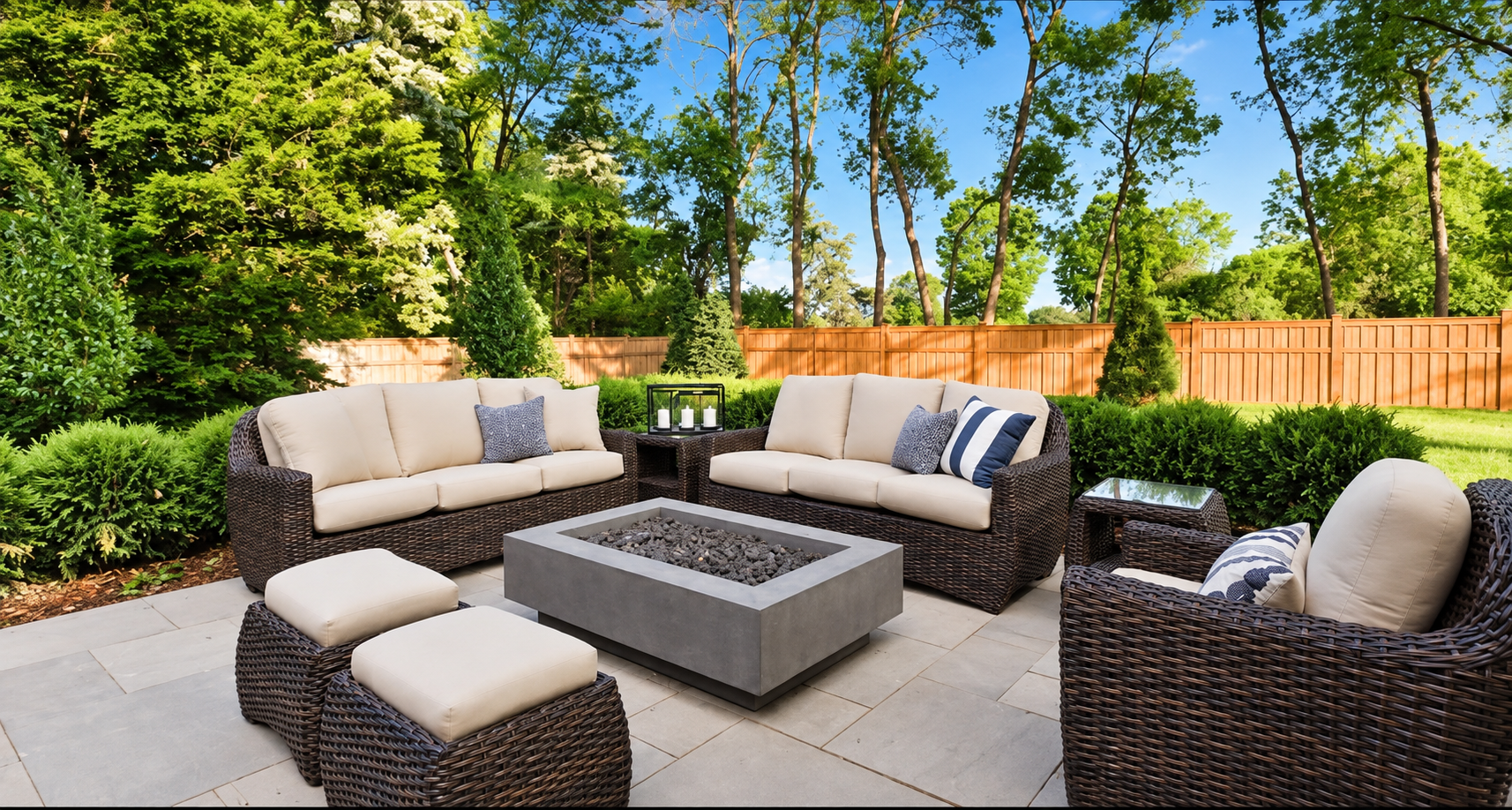 Outdoor patio with wicker sectional seating around a square fire pit, surrounded by trees and a wooden fence.
