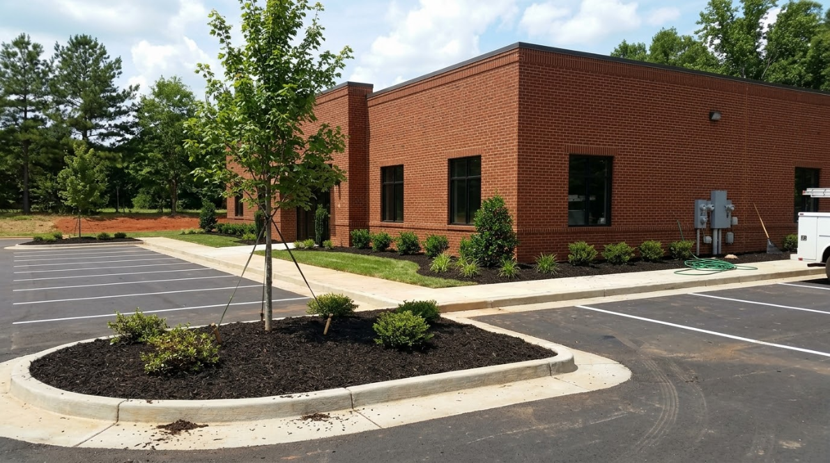 Red brick commercial building with dark windows, landscaped planter, and parking lot on a sunny day