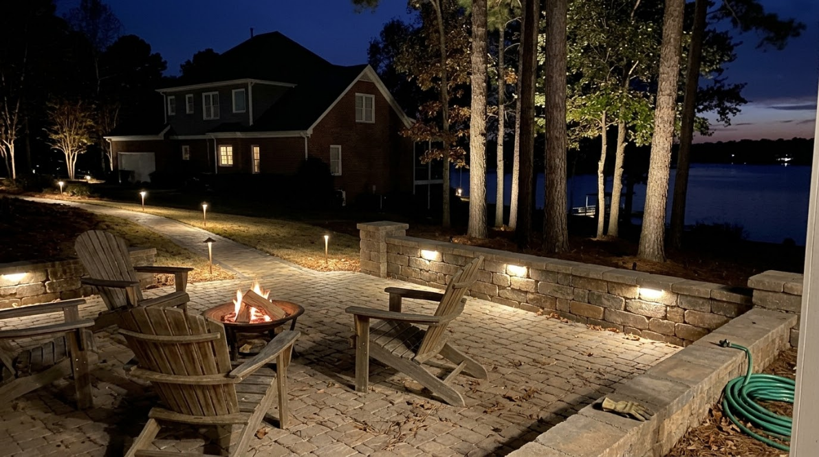 Nighttime lakeside patio with chairs, fire pit, and lit stone wall beside a cabin and trees.