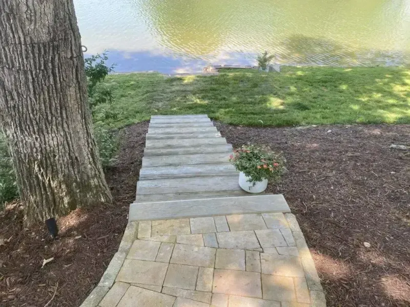 Stone path steps down a grassy slope beside a tree and potted flowers toward the water.