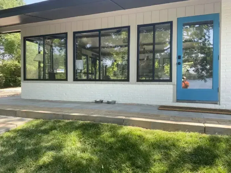 Single-story storefront with large black windows, a blue door, and a white brick exterior beside a grassy lawn