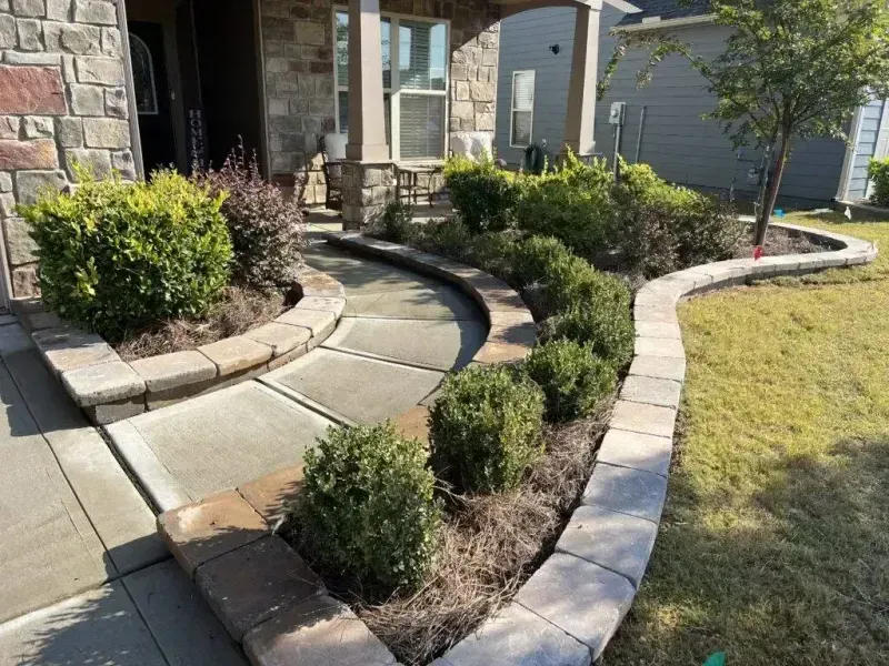Curved stone walkway and garden beds beside a stone house, with trimmed shrubs and a grassy lawn.