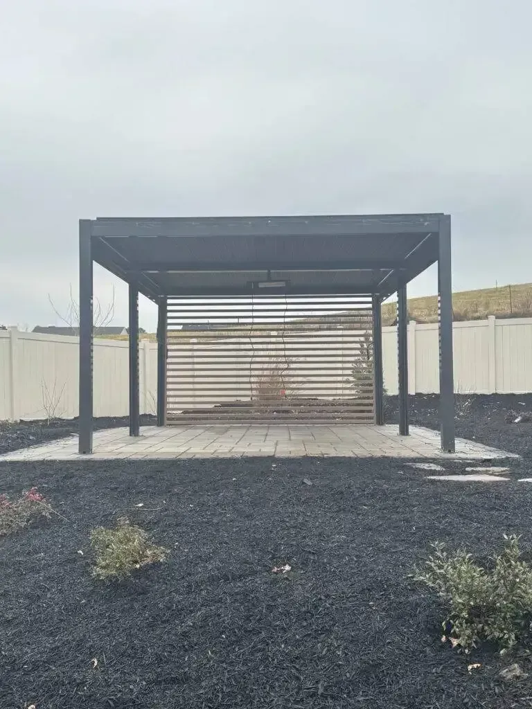 Open metal pavilion with slatted back wall in a gravel yard under a cloudy sky