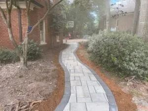 Curved stone walkway through a landscaped garden between brick buildings, lined with shrubs and trees.
