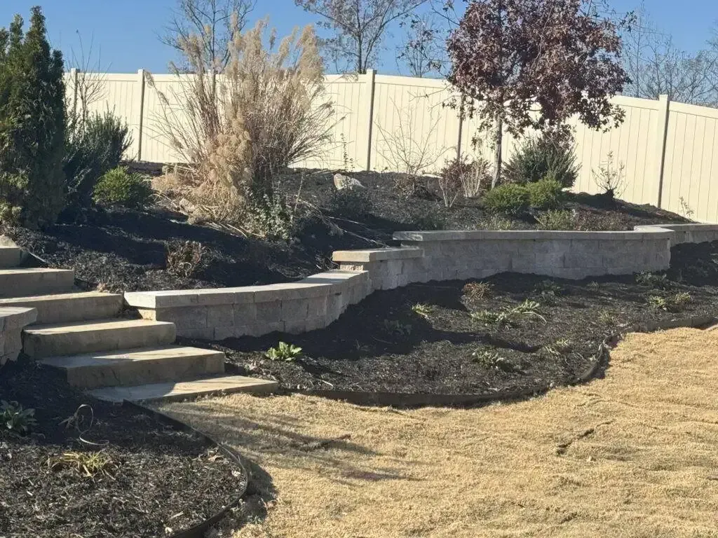 Landscaped backyard with stone retaining walls, shrubs, and a curved mulch path under a bright sky