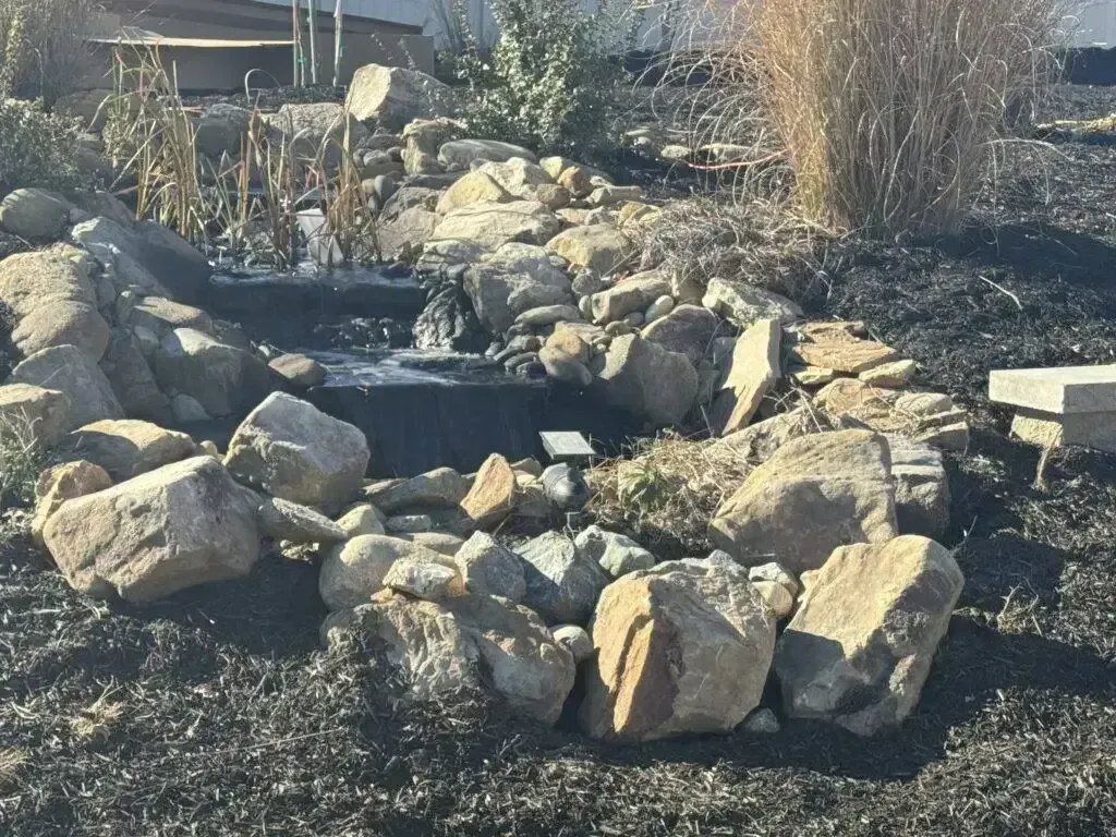 Rock-lined garden pond with a small waterfall, boulders, and dry grasses in sunlight