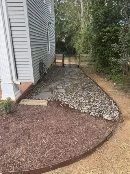 Gravel side yard path beside a gray house, bordered by mulch and leading to a gate at the back.