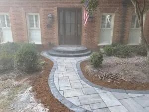 Curved stone walkway leading to a brick front door with steps, bordered by shrubs and mulch.