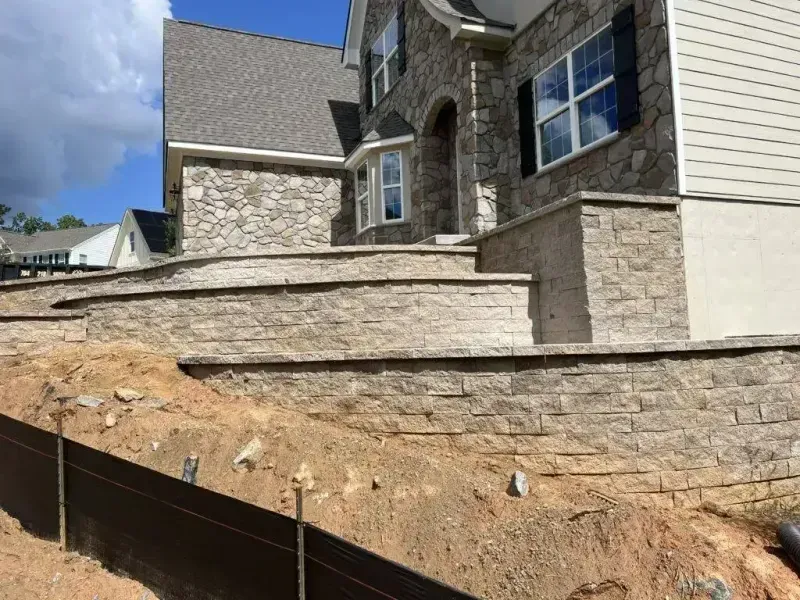 Stone house exterior with retaining wall and sloped dirt yard under a cloudy sky