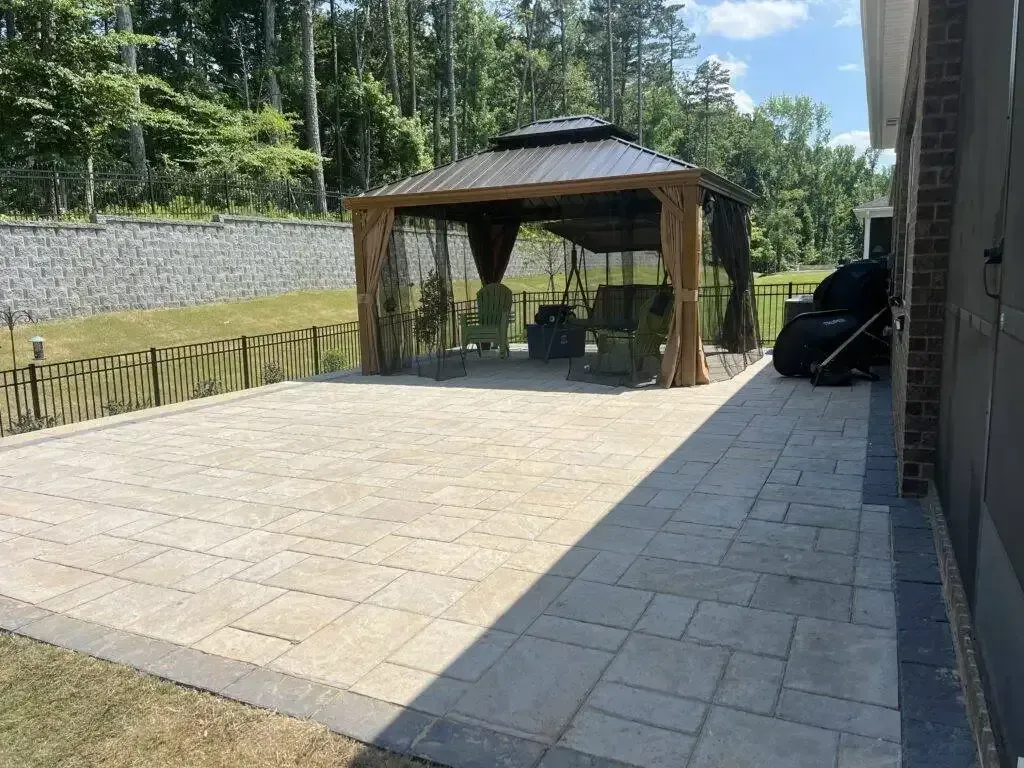Paved patio with a wooden gazebo and chairs beside a house, backed by a stone wall and trees