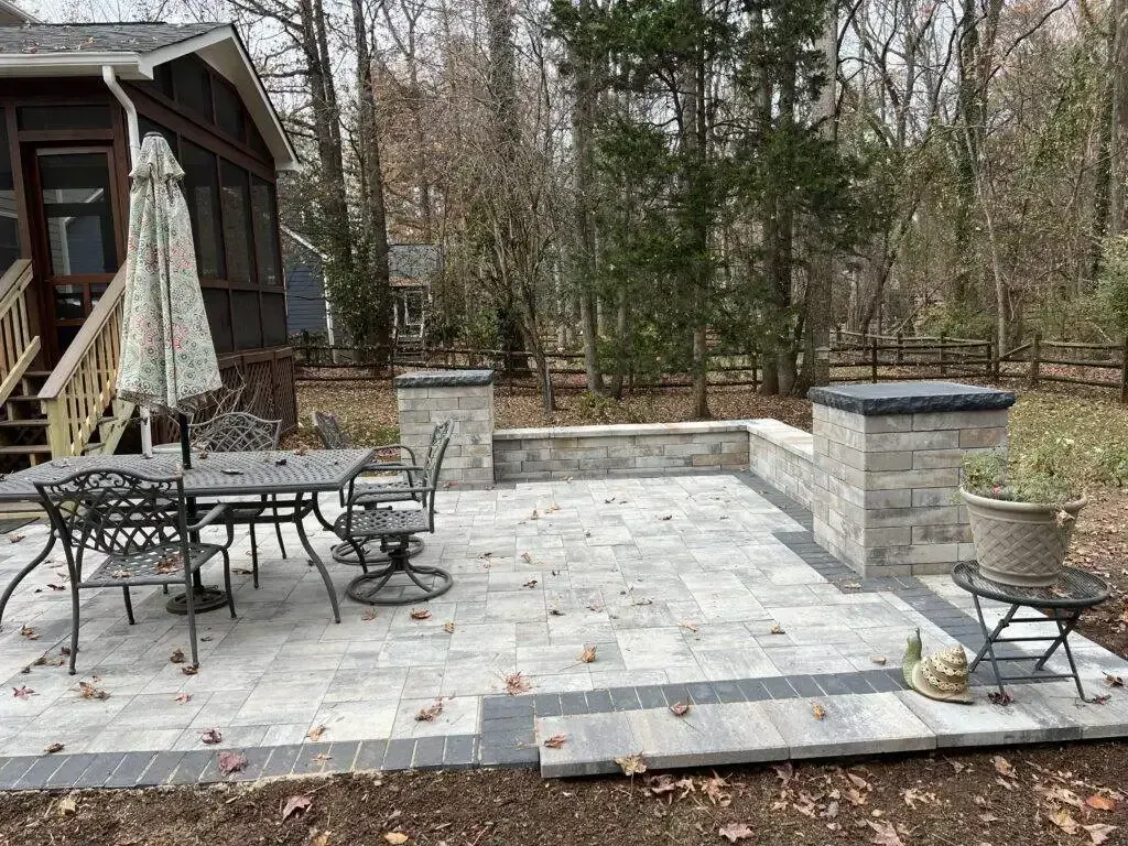 Backyard stone patio with table, chairs, and built-in grill beside a wooded deck area