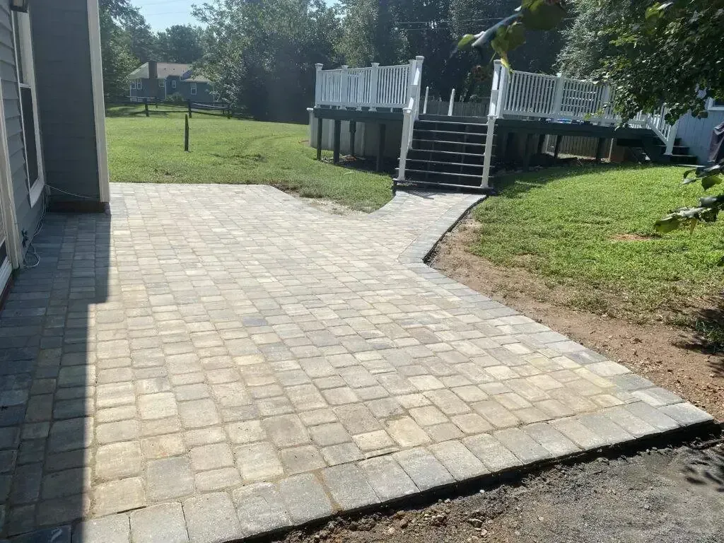 Paver patio beside a house leading to a raised wooden deck with stairs in a grassy yard.