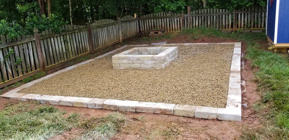 Rectangular gravel yard bed bordered by concrete blocks, with a raised concrete structure in the center and a fence behind.