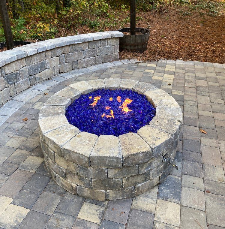 Stone fire pit with blue glass and flames, set in a paved backyard patio beside a curved stone wall