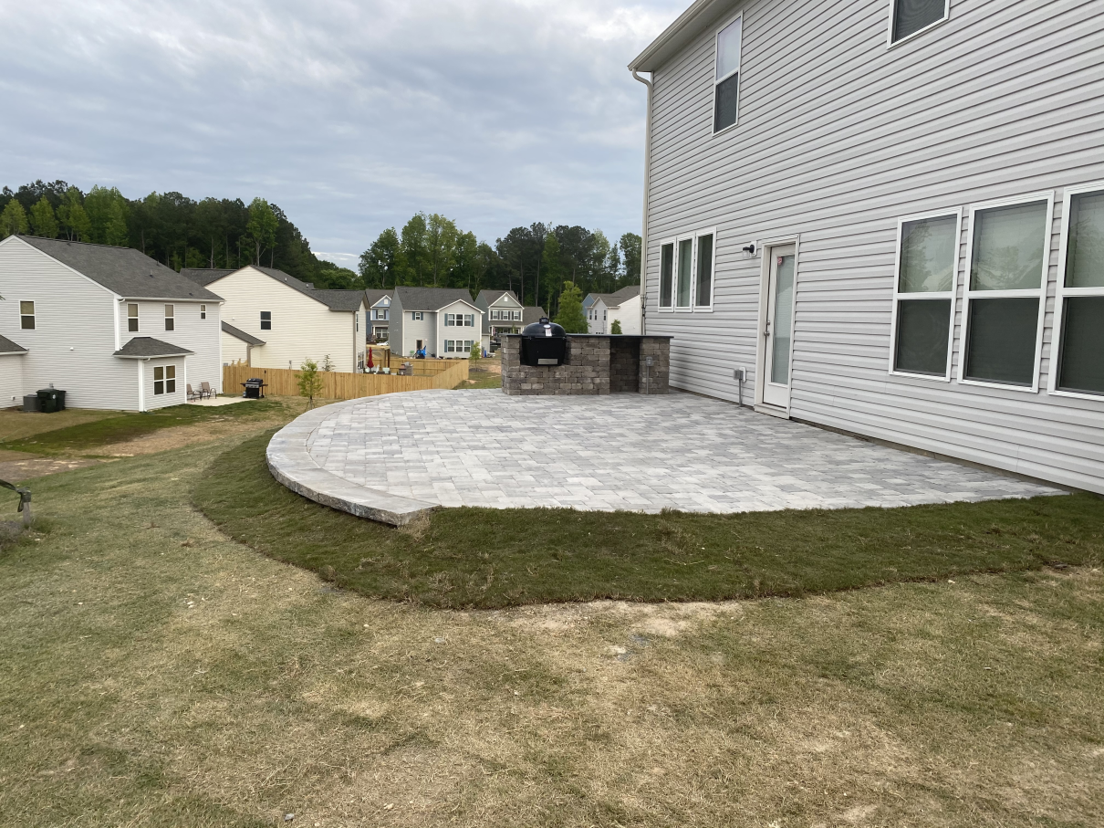 Backyard with a curved stone patio beside a white house and neighboring homes under cloudy skies