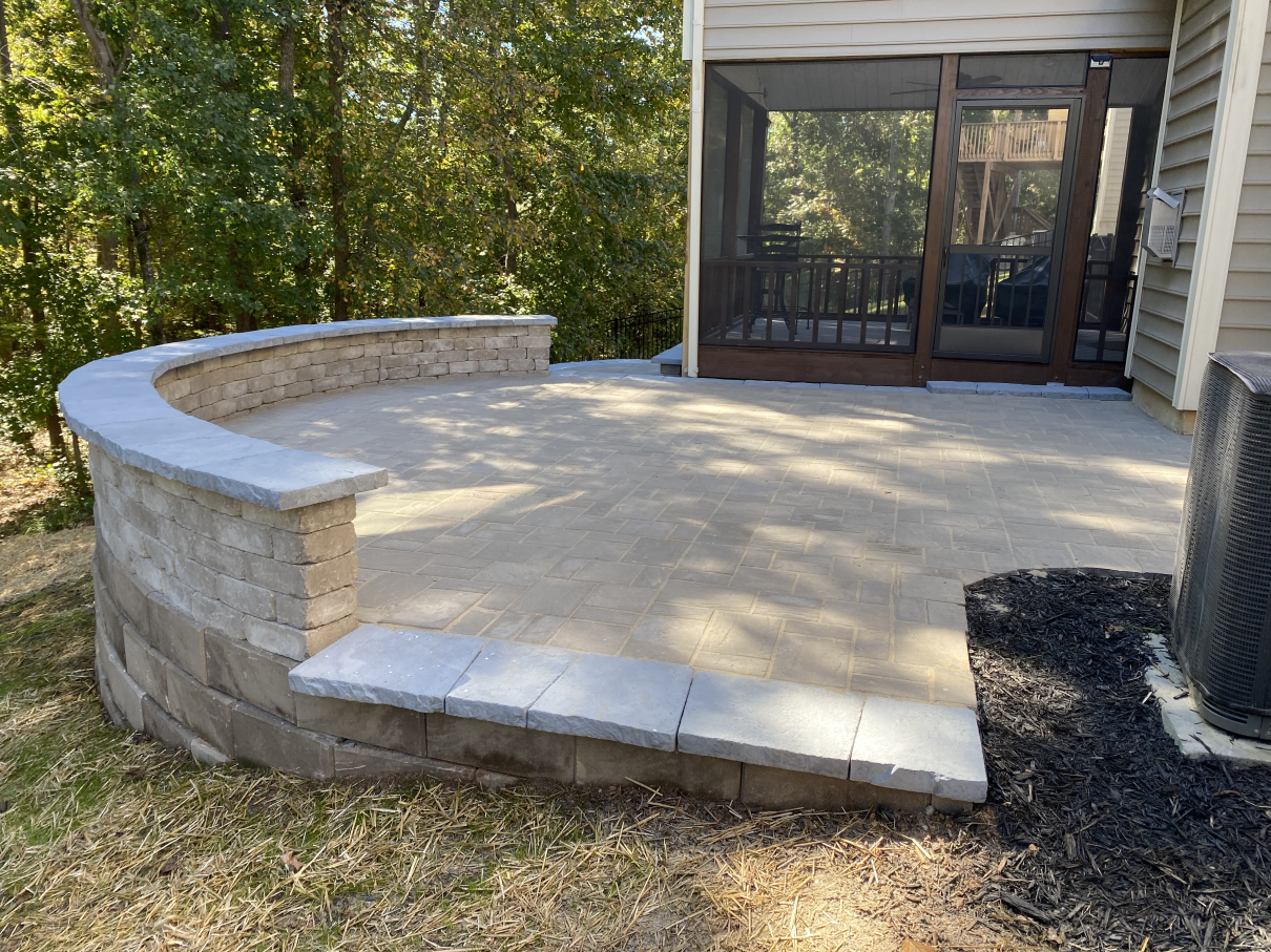 Patio with curved stone retaining wall, concrete floor, and screened porch beside a house and trees
