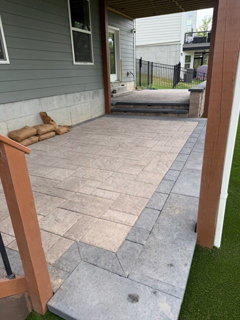 Covered backyard patio with light stone pavers, gray border tiles, and sandbags against the house wall.