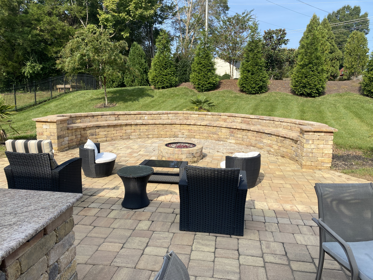 Backyard patio with wicker chairs around a stone fire pit, bordered by a curved stone wall and grassy hill.