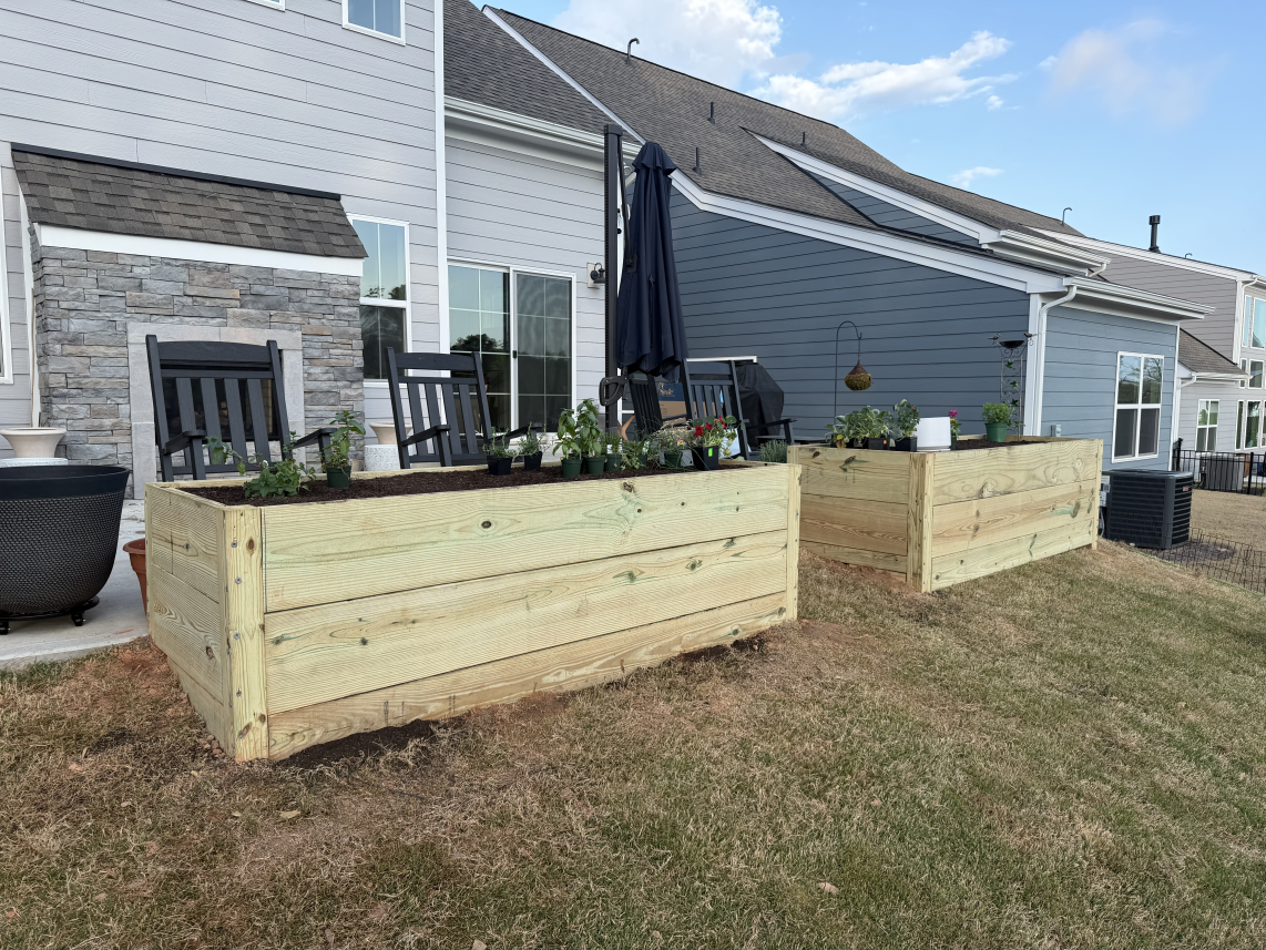 Raised wooden garden beds in front of a house with stone steps and black siding.