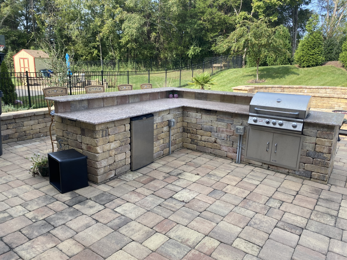 Outdoor stone patio with built-in brick grill, sink, and black fire pit beside a grassy yard