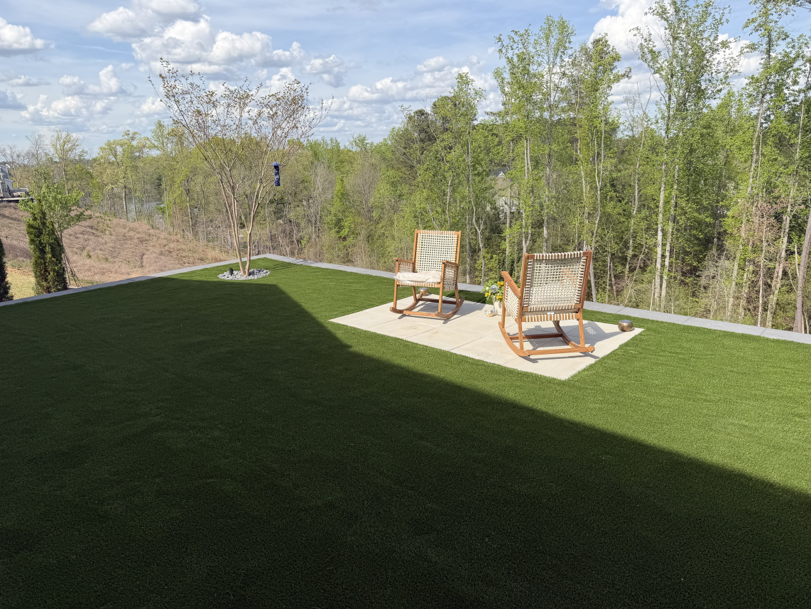 Patio with two wooden chairs on a small deck beside a green lawn and trees under a blue sky