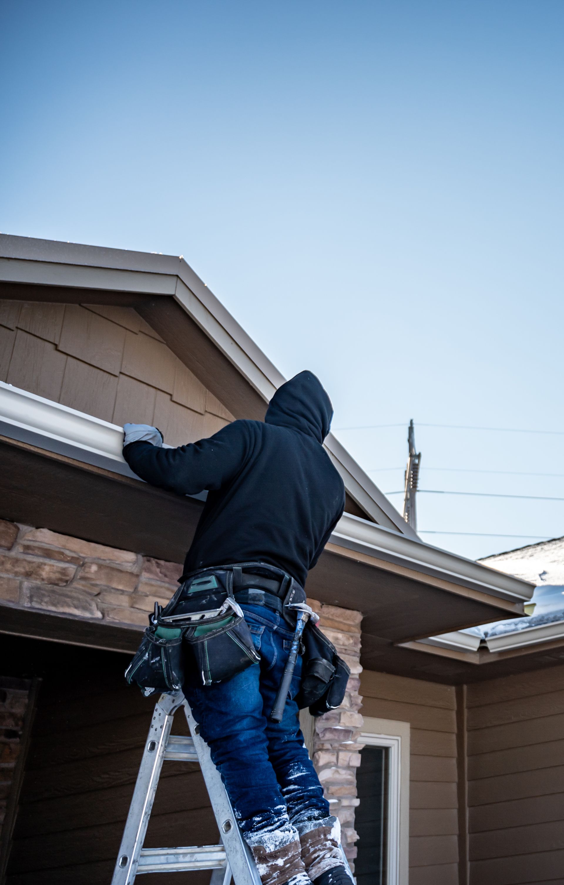 Roofer on a gray shingle roof near a chimney, surrounded by trees and a sunny sky.