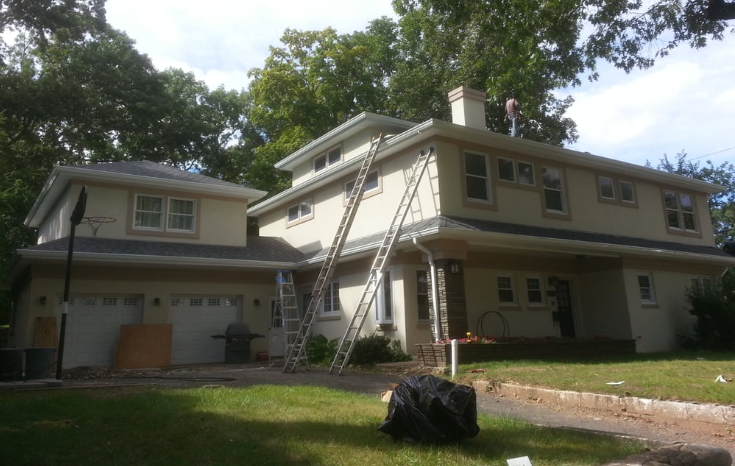 Roofer on a gray shingle roof near a chimney, surrounded by trees and a sunny sky.