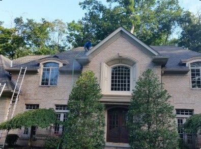 Roofer on a gray shingle roof near a chimney, surrounded by trees and a sunny sky.