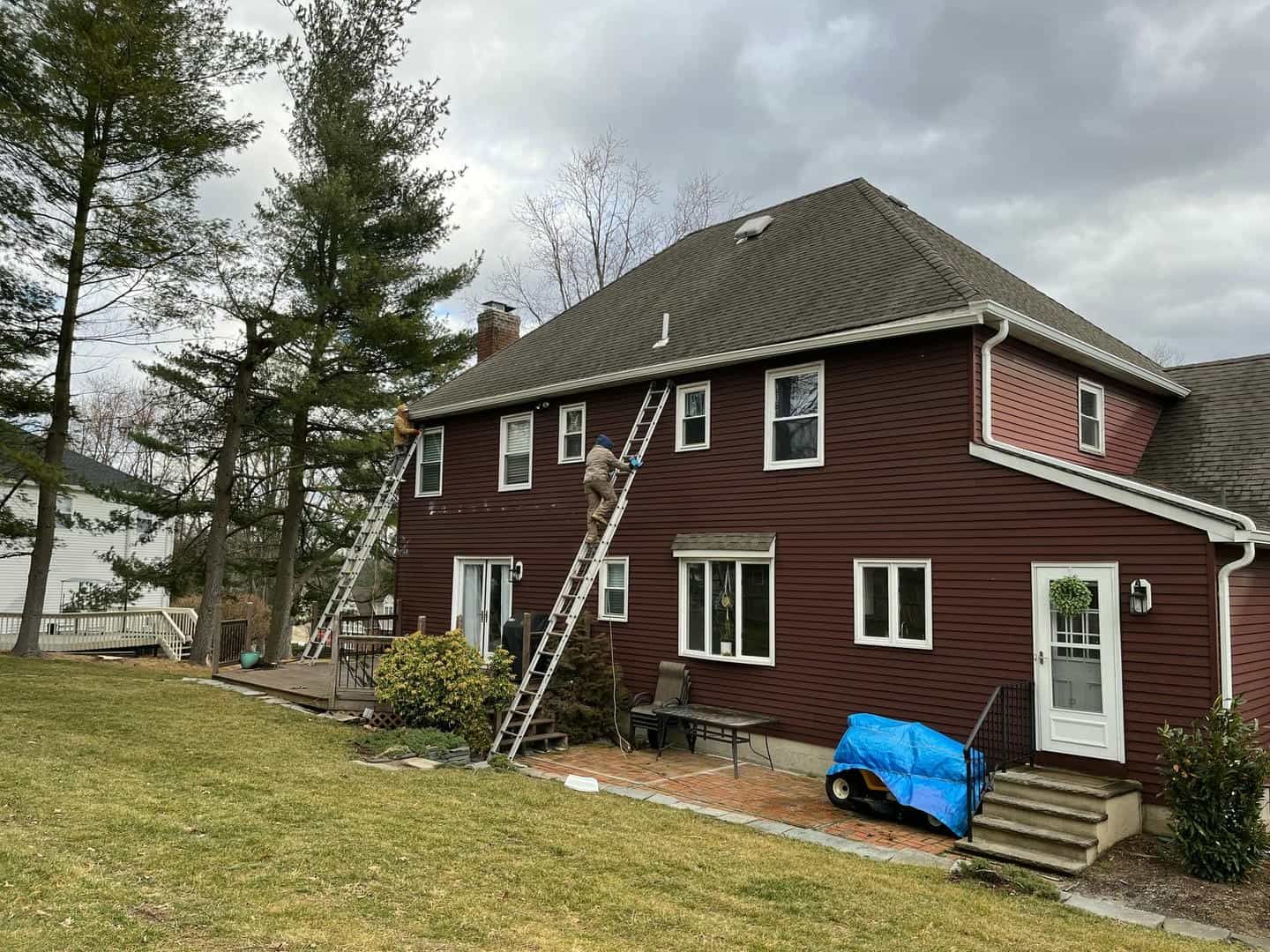 Two ladders leaning against a brown house, person climbing one. Overcast sky.