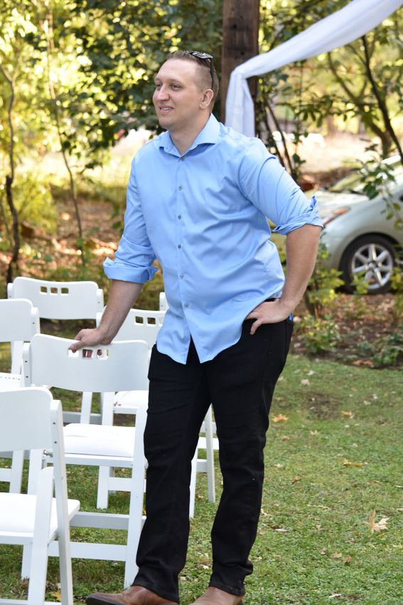 Man in a light blue shirt standing outdoors beside white chairs at a garden event