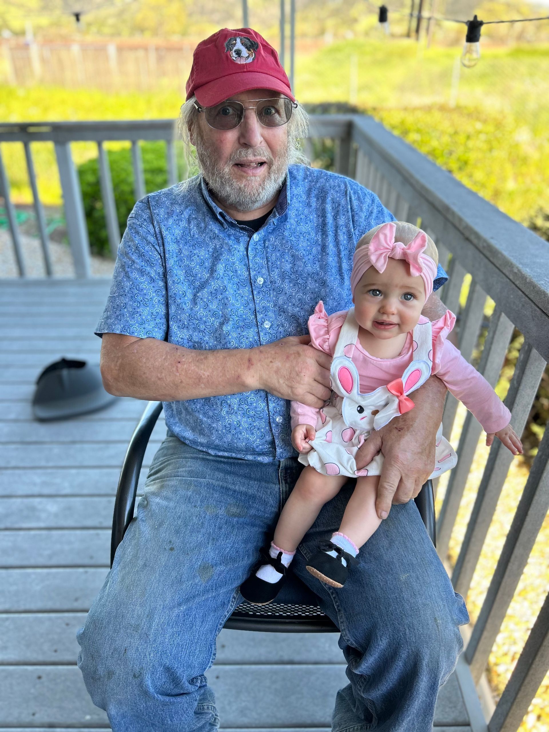 Man in red cap holding a smiling baby on a porch with a railing and grassy background